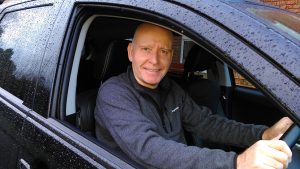 Paul Tracey sits in the driving seat of a parked car. The window is down and he's smiling at the camera.